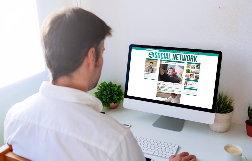un homme est de dos en chemise blanche. il regarde l'écran de son ordinateur sur lequel est affiché une page internet avec écrit en vert "Social Network"