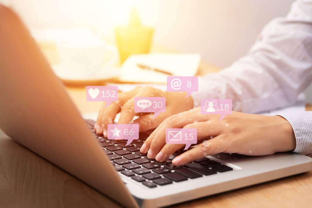 Les mains d'une personne sur le clavier d'un ordinateur portable. La personne est entrain d'interagir sur les réseaux sociaux. Divers icônes (likes, favoris, message, communauté, tags) de couleur violet flottent au dessus du clavier.