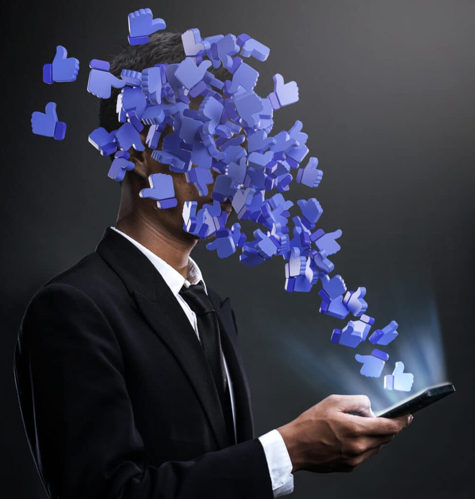 Un homme portant un costume et une cravate tient dans sa main un Smartphone d'où sortent des pouces bleus levés en guise de "likes" qui lui couvrent le visage.