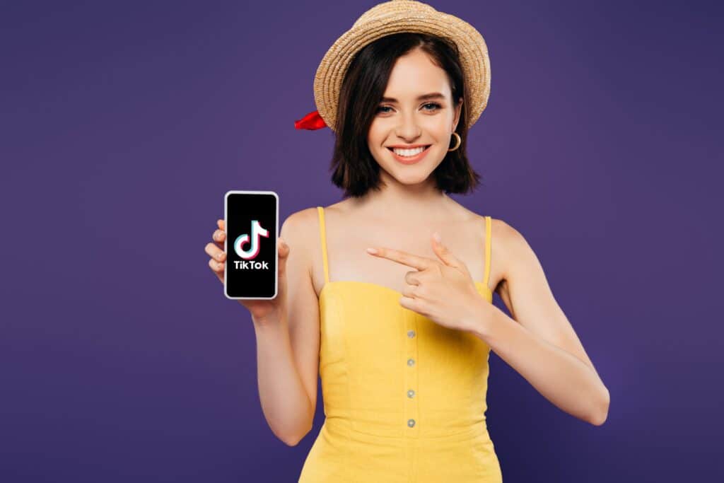 Une jeune fille souriante montrant du doigt un smartphone qu'elle tient dans l'autre main, sur lequel est affiché le logo de TikTok